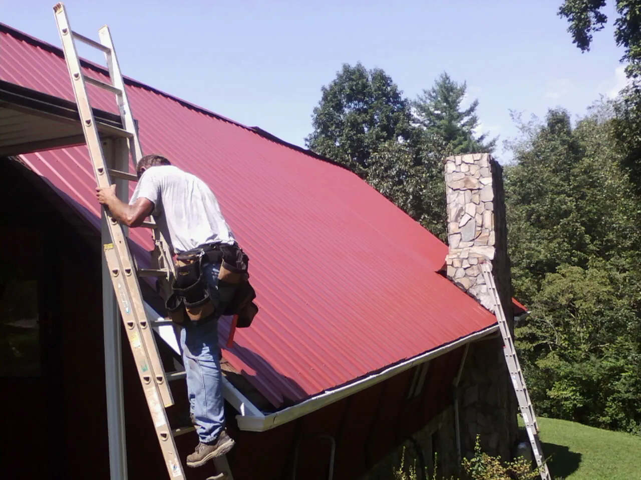 Roofer on a ladder beside a red metal roof and stone chimney — Soffit & Fascia Repair by Roofing Brothers in Kerrville